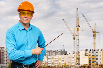 Builder worker in a hardhat standing and showing with pointer stick on construction site with cranes behind of him.