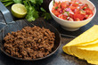 fried ground beef in a pan, as a filling for taco shells with ingredients as tomato salsa, lime and herbs on a dark stone background
