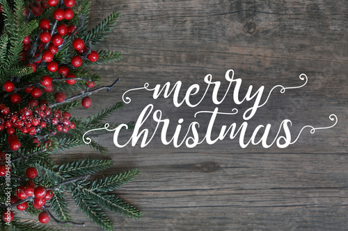 Fotografie, Obraz Merry Christmas Text with Evergreen Branches and Berries Over Dark Rustic Wood B