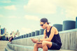 © Alexander Image - Man travels in New York in hot summer. Wearing black tank top, shorts, cap worn backward, young guy sits on wooden stairs at park, reads messages on cell phone. Filtered effect..