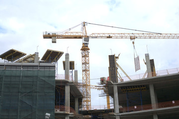 Tower hoisting crane lifts panels at construction process on top of constructed building over sky with clouds