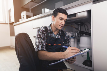 The plumber sits next to the kitchen sink on the floor and writes a report on the work done.