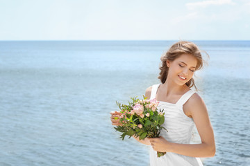 Wall Mural - Young smiling bride in white gown holding bouquet on seashore