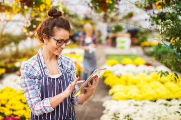 Portrait of professional modern florist business smiling girl with eyeglasses and tablet in the greenhouse full of flowers and pots searching for improvement on the internet.