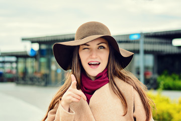 Poster - Close-up portrait of girl in a brown hat and beige coat eye winks at the camera, points the finger at you and smiles. Choose you