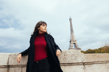 Full length portrait of young woman in black trench coat standing on embankment near Eiffel tower in Paris, France