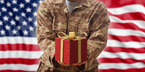 Veterans Christmas gift box.  American soldier in a military uniform offering a red package on US flag background