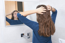 Beautiful Teenage Girl Doing Her Hair Before The Mirror, Standing In A Bathroom And Looking At Her Reflection In A Mirror