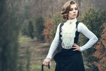 Portrait of gorgeous young woman with elegant Victorian hairstyle wearing old-fashioned gown with jabot leaning on her cane and looking aside, misty autumn park on background. Vintage style