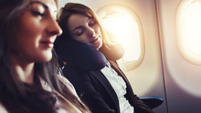 Girlfriends Traveling By Plane. A Female Passenger Sleeping On Neck Cushion In Airplane.