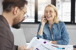 © WHstudio Leushin N - Portrait of successful young smiling businesswoman has cheerful expression, talks on mobile phone while her partner studies documents, meet for preparing to conference. Two colleagues collaborate