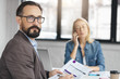 © wayhome.studio  - Serious bearded man wears formal suit, works with documents, reads contract and female model in background speaks on smart phone. Businessman at office desk, analyzes investment chart and turnover