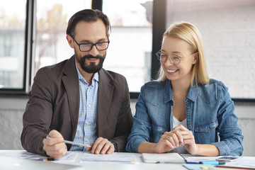 Happy coworkers sit at working desk, look down in papers, point out financial data on report, check balance, analyze statistics, have happy expressions. People, business, career, job concept