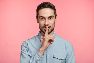 Wall Mural - Attractive pleasant looking unshaven man keeps finger on lips, asks friends not to tell secret and what surprise he prepared for girlfriend, shows hush sign, gestures at studio. Silence and people