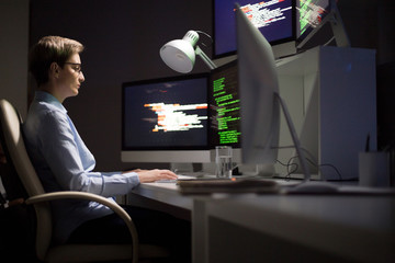 Portrait shot of concentrated middle-aged software developer wrapped up in work while sitting in front of computer at dim office