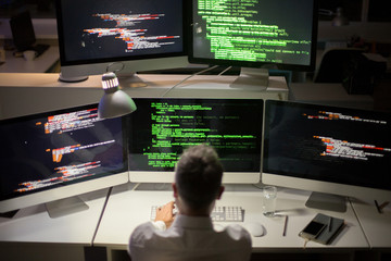 Unrecognizable gray-haired software developer writing code while sitting in front of modern computer, interior of dim office on background
