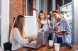© undrey - Young female manager sitting at desk pointing at laptop explaining giving tasks to her employees standing writing the information in notebooks in office.