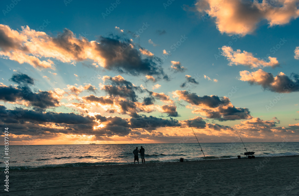 silhouettes of two men fishing on the shore as morning sunlight breaks ...