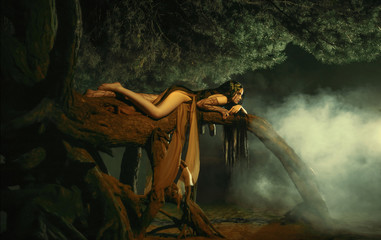 A fabulous, forest nymph with long hair lies on a tree branch with an aggressive look. Background dark night and fog. Mythical character of Gian