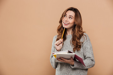 Wall Mural - Portrait of happy thinking female student in warm sweater holding pencil and notebooks, looking aside
