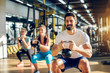 Attractive handsome bearded man holding kettlebell and doing squats in a fitness group with two girls in the modern gym.