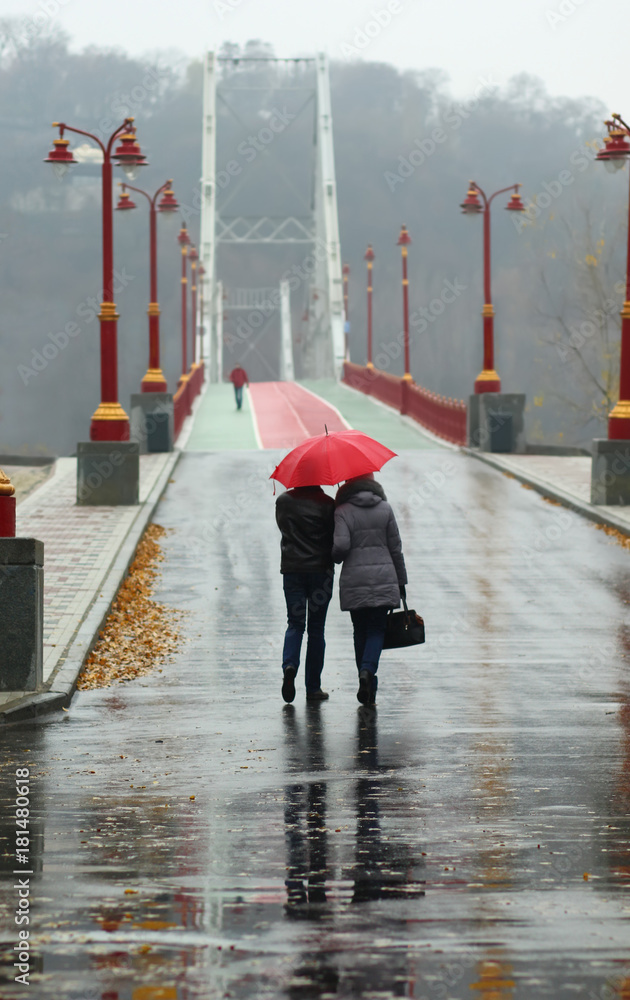 Couple walking under red umbrella on a rainy foggy day on the downtown Kyiv pedestrian city bridge, back view - obrazy, fototapety, plakaty 