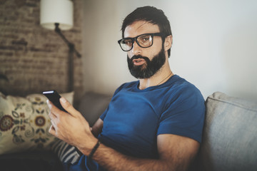 Wall Mural - Young bearded man wear casual clothes and eye glasses typing fingers on smartphone screen at modern loft home.Horizontal.Blurred background.