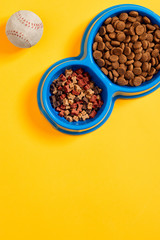 Dry pet food in bowl with a ball on yellow background top view