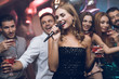 © VadimGuzhva - A woman in a black dress is singing songs with her friends at a karaoke club. Her friends have fun on the background.
