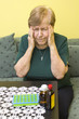 Starsza kobieta trzyma się za skronie z grymasem bólu na twarzy. Pojemniki z lekami oraz buteleczki leżą na stole obok niej.