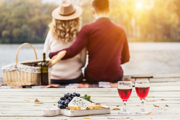 Romantic dinner outdoors. Glasses with red wine, grapes and cheese on wooden board. Young couple having romantic dinner outdoors. Back view man hugging her beautiful girlfriend. Couple in love.