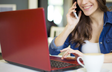 We can help you. Beautiful smiling brunette women sitting at the table and working at a laptop and speaking on smartphone. Co working zone. Pretty freelancer.