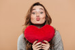 Close-up photo of lovely brunette girl holding big soft red heart while sending kiss, looking at camera