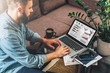 © foxyburrow - Young bearded hipster man, entrepreneur sits on couch at coffee table,uses laptop with graphs,charts, diagrams on screen.On table notebook,cup of coffee.Online marketing,education,e-learning. Startup.