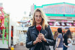 © BublikHaus - Portrait of cute and beautiful young blonde woman in trendy outfit, looking into camera at county fair or festival with sweet candy apple covered in caramel, at holiday vacation event