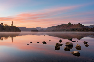 Beautiful pink sunrise reflected in a calm misty lake with rocks in foreground. Taken at Derwentwater in the English Lake District.