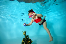 Little Girl Underwater In The Pool Hangs A Toy On The Christmas Tree. Portrait. Shooting Under Water. Horizontal Orientation