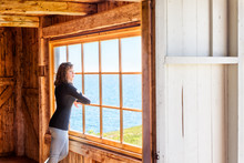 Side Of One, Lonely, Alone Young Woman Standing By Large Glass Window Looking At Peaceful Ocean View, Cliff, In Rustic Wooden House