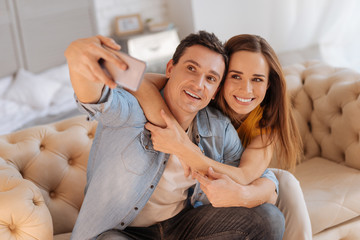 Nice photo. Cheerful beautiful loving people sitting together on a comfortable sofa and looking at the screen of a smart phone while taking photos