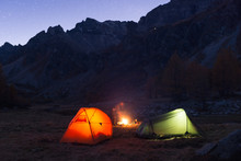 Friends Camping Under Starry Sky With Two Tents And Campfire In Blue Hour At Night.
