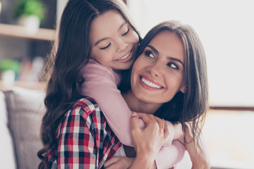 Poster - Playful small girl with long dark hair is hugging her mum's neck and looking at her with a smile, they have perfect weekends at home