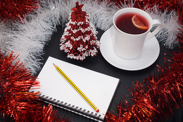 Wall Mural - Cup of red tea with lemon slice, notebook with pencil, red and white christmas decoration and red christmas tree on dark table