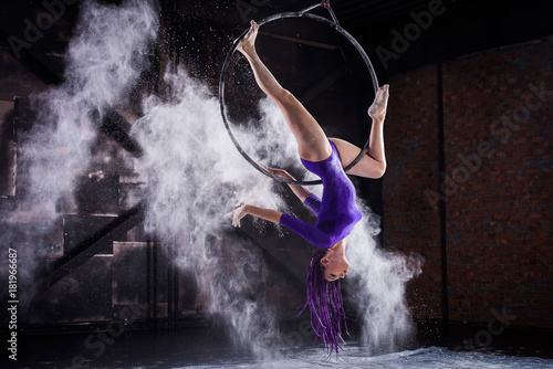 Fotografía A young, slender girl in a lavender leotard and lilac dreads, doing gymnastics on the air ring, the Hoop and throws the hands of the white powder in the form of a cloud