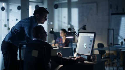 Late at Night Two Industrial Engineers Discuss 3D Component Design Made with CAD Program on Personal Computer. Office Looks Modern with Lots of Blueprints on the Walls.