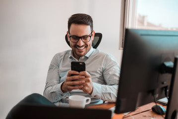 Portrait of a happy smiling businessman in eyeglasses using smartphone while sitting at the office.