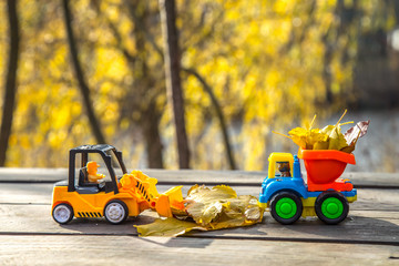 Two small toy truck and tractor is loaded with yellow fallen leaves. The car stands on a wooden surface against a background of a blurry autumn park. Cleaning and removal of fallen leaves. Seasonal wo