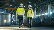 © Gorodenkoff - Back View Shot of Male and Female Industrial Engineers Having Discussion While Walking Through Heavy Industry Manufacturing Factory. Big Metalwork Constructions, Pipeline Elements Lying Around.