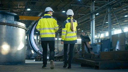 Wall Mural - Back View Shot of Male and Female Industrial Engineers Having Discussion While Walking Through Heavy Industry Manufacturing Factory. Big Metalwork Constructions, Pipeline Elements Lying Around.