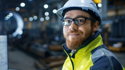  Industrial Engineer Wearing Hard Hat,  Safety Jacket and Glasses Smiles on Camera. He Works in Big Heavy Industry Factory.