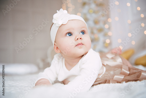 Baby girl wearing cute dress and headband, lies on a white cover in festively decorated room with garland of lights. With surprise watches in the camera, on a background a set of bright fires.